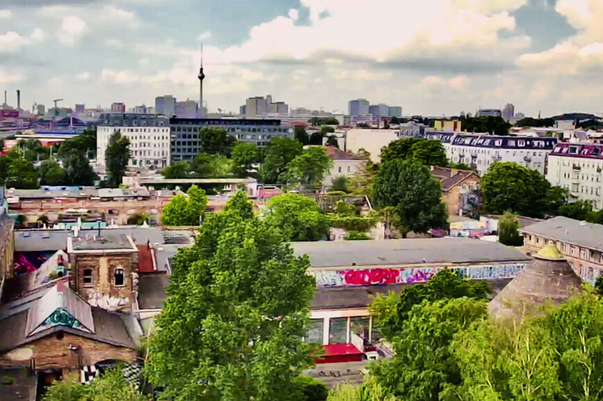 Das RAW Gelände in Berlin-Friedrichshain aus der Vogelperspektive. Rechts unten ragt die Spitze des Kletterturms "Der Kegel" aus dem Grün der Bäume hervor und am Horizont ist der Fernsehturm zu erkennen. 